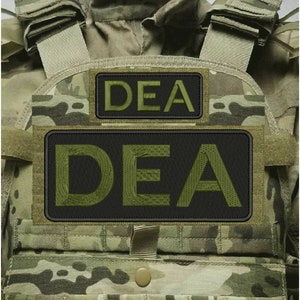 May include: A tactical vest featuring a camouflage pattern in shades of green, brown, and tan. The vest includes two rectangular black patches with embroidered olive green text that reads DEA, positioned on the front chest area.