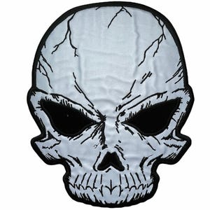May include: A light gray skull patch with a black embroidered border. The skull features deep black eye sockets, a small triangular nose cavity, and detailed black line work depicting cracks and facial contours across the surface.