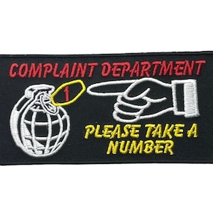 May include: A rectangular black embroidered patch featuring the red text COMPLAINT DEPARTMENT at the top. Below, a white hand points toward a white grenade with a yellow circle containing a red number one. Yellow text reads PLEASE TAKE A NUMBER.