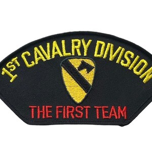 May include: A black embroidered patch with a curved top edge. The text 1ST CAVALRY DIVISION is stitched in yellow thread at the top. A yellow shield with a black diagonal stripe is in the center. The text THE FIRST TEAM is stitched in red below.