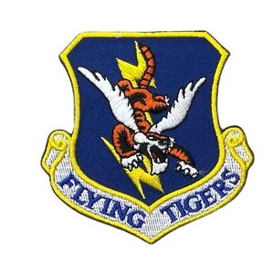 May include: A shield-shaped embroidered patch featuring a blue background with a yellow border. The design depicts an orange and black winged tiger leaping over a yellow lightning bolt. Below the tiger, a white scroll displays the text FLYING TIGERS.
