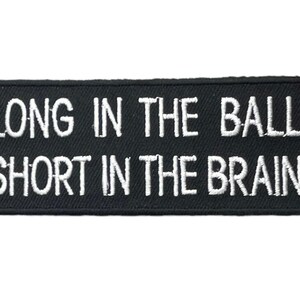 May include: A rectangular black embroidered patch with rounded corners featuring the white stitched text LONG IN THE BALLS SHORT IN THE BRAINS arranged in two lines. The patch has a thick black border and a textured fabric surface.