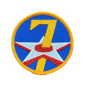 May include: A circular embroidered patch featuring a bright blue background with a yellow number 7 centered over a white five-pointed star. The star has a red circular center and red outlines, all enclosed within a thick yellow stitched border.