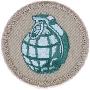 May include: A circular embroidered patch featuring a light beige background with a dark beige stitched border. Centered on the patch is a detailed thread-stitched graphic of a hand grenade in shades of light blue and dark green.