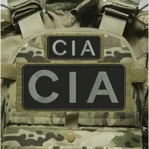 Puede incluir: Un chaleco táctico de camuflaje con dos parches rectangulares negros con el texto CIA bordado en gris. El chaleco incluye correas para los hombros, correas de sujeción y un bolsillo frontal con un cierre de botón central en camuflaje.