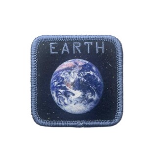 May include: A square embroidered patch featuring a dark blue background with a depiction of the Earth in the center. The word EARTH is stitched in light blue capital letters above the planet. The patch has a light blue stitched border.