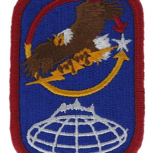 May include: A rectangular embroidered patch with a red border on a blue background. It features a brown eagle with a white head, a yellow lightning bolt, a white star, a red arrow, and a white globe graphic at the bottom with a mountain silhouette.