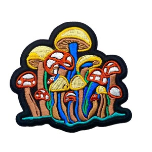 May include: An embroidered patch featuring a cluster of mushrooms with yellow, orange, and white caps, tan stalks, and vibrant blue stems. The design is outlined in black thread with a teal base, creating a detailed, multi-colored botanical motif.