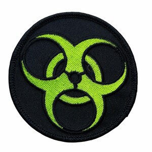 May include: A circular embroidered patch featuring a bright neon green biohazard symbol centered on a solid black fabric background. The patch has a thick, stitched black border around the entire circumference, creating a clean, finished edge.