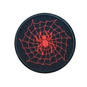 May include: A circular embroidered patch featuring a black background with a red spider centered in the middle of a red stitched spider web. The patch has a thick black border around the entire circumference, creating a distinct circular shape.