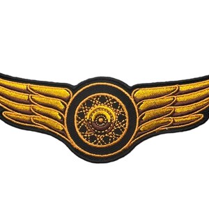 May include: An embroidered patch featuring a central circular wheel design with intricate gold and brown stitching, flanked by two symmetrical golden wings extending outward, all outlined with a thick black border on a black fabric base.