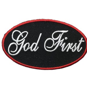 May include: An oval-shaped embroidered patch with a black background and a red border. The text God First is stitched in white cursive lettering across the center of the patch. The design features a clean, bold contrast between the colors.