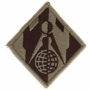 May include: A diamond-shaped embroidered patch featuring a two-tone brown and tan design. The graphic depicts a stylized compass or drafting tool positioned above a globe, with the colors split vertically across the entire emblem and border.