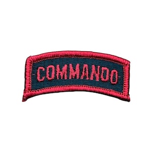 May include: A curved, rectangular embroidered patch featuring a dark blue background with a red border and the word COMMANDO stitched in red capital letters across the center. The patch has a distinct arched shape and a textured fabric finish.