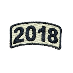 May include: A curved, rectangular embroidered patch featuring the black stitched numbers 2018 centered on a cream-colored background. The patch is bordered by a thick black stitched edge, creating a distinct outline for the numerical design.