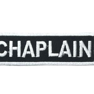 May include: A rectangular embroidered patch with a black background and a white border. The word CHAPLAIN is stitched in bold, white, capital letters across the center of the patch. The design features a clean, high-contrast aesthetic.
