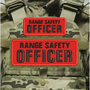 May include: A camouflage tactical vest featuring two rectangular red patches with black embroidered text that reads RANGE SAFETY OFFICER. The patches are positioned horizontally on the front of the vest, which includes shoulder straps and a central snap button.