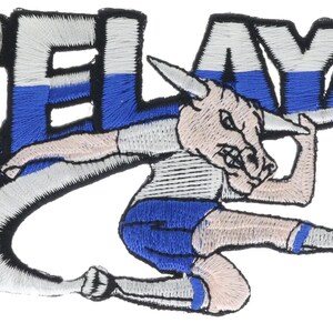 May include: An embroidered patch featuring the word CELAYA in white and blue block letters. A cartoon bull character with a tan face and horns is depicted in a dynamic pose, wearing a white shirt and blue shorts while kicking a black and white soccer ball.