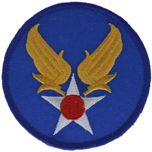 May include: A circular embroidered patch featuring a blue background with a central white five-pointed star containing a red circle. Above the star are two symmetrical golden yellow wings with detailed stitching, all enclosed by a blue border.
