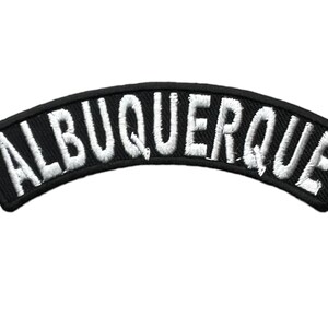 May include: A curved, black embroidered patch featuring the word ALBUQUERQUE in bold, white, capitalized, sans-serif lettering. The patch has a thick black border and a textured fabric finish, designed to be applied to clothing or other textile items.