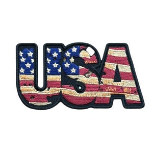 May include: An embroidered patch in the shape of the letters USA. The design features the American flag pattern with a blue field containing white stars on the left and horizontal red and white stripes throughout, all outlined in black thread.
