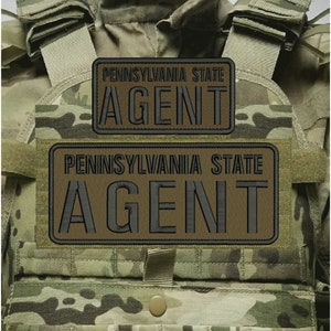 Può includere: Un gilet tattico mimetico con due toppe rettangolari con testo ricamato in nero che recita PENNSYLVANIA STATE AGENT. Il gilet include spallacci regolabili, una chiusura centrale con bottone a pressione e un motivo multicolore.