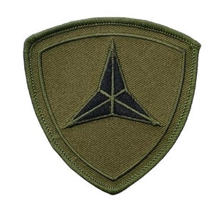 May include: A shield-shaped embroidered patch in olive green featuring a dark navy blue three-pointed star emblem centered within the fabric. The patch has a thick, stitched border and a textured twill weave finish throughout the entire surface.