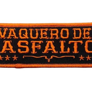May include: A rectangular embroidered patch with a black background and a vibrant orange border. The text VAQUERO DEL ASFALTO is stitched in orange across the center, flanked by three small orange stars on each side and a decorative line below.