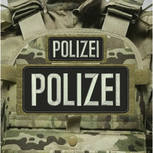 Op de afbeelding: Een tactisch vest met een camouflagepatroon in tinten groen, bruin en beige. Aan de voorzijde zijn twee rechthoekige zwarte patches bevestigd, elk geborduurd met de witte tekst POLIZEI in een vet schreefloos lettertype met een gestikte rand.