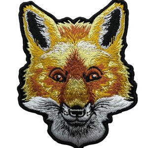 May include: An embroidered patch featuring the head of a fox with orange, yellow, and white fur. The design includes black accents on the ears and around the eyes, with a detailed snout and whiskers, all outlined by a thick black border.