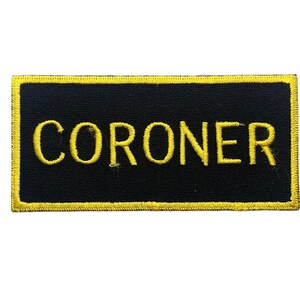 May include: A rectangular embroidered patch with a black background and a yellow border. The word CORONER is stitched in bold yellow capital letters across the center of the patch. The design features a clean, uniform stitch pattern throughout.
