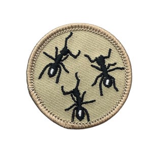 May include: A circular embroidered patch featuring a tan background with a stitched tan border. The design displays three black ants with white highlights on their abdomens, arranged in a scattered pattern across the surface of the patch.