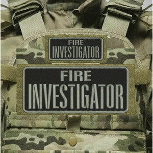 May include: A tactical vest featuring a camouflage pattern in shades of green, brown, and tan. Two rectangular black patches are attached to the front, each embroidered with the white text FIRE INVESTIGATOR in a bold, sans-serif font.