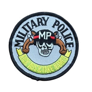 May include: A circular embroidered patch with a light blue background and a black border. It features the text MILITARY POLICE in black, a central skull wearing a helmet with MP and a red stripe, two brown pistols, and a green banner reading KICK ASS & TAKE NAMES.