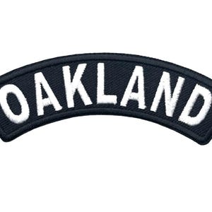 May include: A curved, black embroidered patch featuring the word OAKLAND in bold, white, capitalized, sans-serif lettering. The patch has a thick, stitched black border and a textured fabric surface, designed for application to clothing or gear.