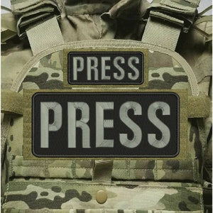 May include: A tactical vest featuring a camouflage pattern in shades of green, brown, and tan. Two rectangular black patches are attached to the front, each embroidered with the word PRESS in large, light-colored, block capital letters.