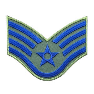 May include: A chevron-shaped embroidered patch featuring a sage green background with three horizontal blue stripes and a central blue star with a circular cutout in the middle. The entire patch is bordered by a light green stitched edge.