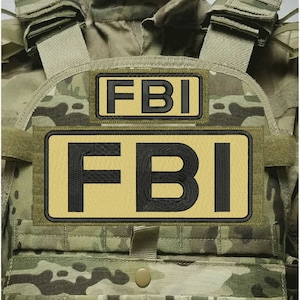 Könnte beinhalten: Eine taktische Weste mit einem Tarnmuster in Grün-, Braun- und Beigetönen. Auf der Vorderseite sind zwei rechteckige Aufnäher angebracht, die jeweils den Text FBI in fetten schwarzen Großbuchstaben auf beigem Grund mit schwarzem Rand zeigen.