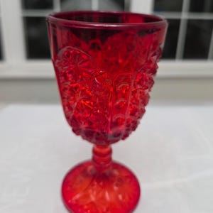 May include: A vibrant red glass goblet featuring a textured hobnail and starburst pattern across the bowl and base. The stem is short and cylindrical, connecting the wide, patterned bowl to a circular footed base with matching embossed details.
