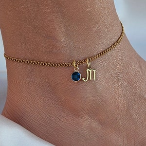 Puede incluir: Cadena de tobillo dorada con una piedra preciosa azul y un dije con la letra "M" colgando.