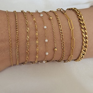 May include: A set of eight gold bracelets, including a chain link bracelet, a curb chain bracelet, a rope chain bracelet, a figaro chain bracelet, a delicate chain bracelet with small pearls, and a simple chain bracelet. The bracelets are stacked on a person's wrist.