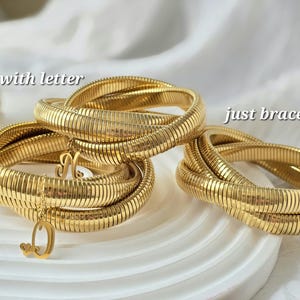 May include: Two gold snake chain bracelets, one with a letter charm, the other without.  The bracelets are layered on a white surface.