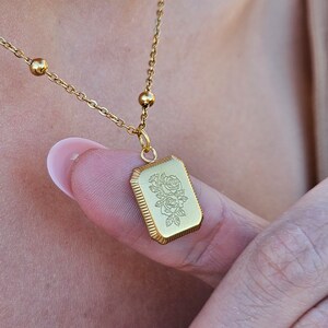 Puede incluir: Un collar de cadena dorada con un colgante rectangular con un diseño de rosa grabado.
