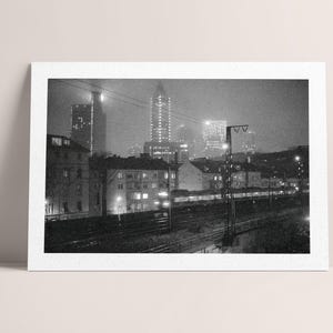 Könnte beinhalten: Schwarz-Weiß-Fotografie einer nächtlichen Stadtlandschaft. Hohe Gebäude mit beleuchteten Fenstern und Lichtern sind im Hintergrund zu sehen. Ein Zug fährt auf Gleisen im Vordergrund, mit einem bewölkten Himmel darüber.