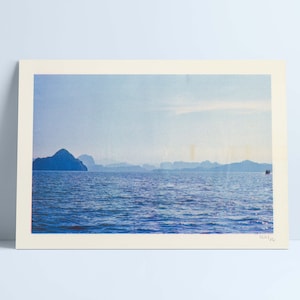 May include: A rectangular landscape print featuring a blue ocean with gentle waves under a bright sky. In the distance, silhouettes of mountains rise above the horizon line. A small boat is visible on the right side. The print has a cream border.