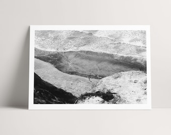 Könnte beinhalten: Eine schwarz-weiße Landschaftsaufnahme, die eine Ansicht einer felsigen Küste von oben zeigt. Das Bild fängt Wellen ein, die gegen das Ufer schlagen, sowie strukturierte Gesteinsformationen und eine kleine, einsame Figur am Strand.