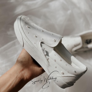 Könnte beinhalten: Weiße Slip-On-Sneaker mit verstreuten Perlen verziert. Die Schuhe haben ein klassisches Design mit einer Gummisohle und elastischen Seitenteilen. Der Text "Sparkly Wedding Co." ist auf der Seite des Schuhs zu sehen.