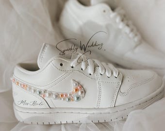 May include: White leather low-top sneakers with a pearl and iridescent embellishment on the side. The sneakers have a white swoosh logo and the text "Mrs. Bride" is written on the side.