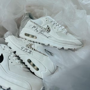 May include: White sneakers with crystal embellishments and the text "Mrs Thorund" and "4.25.26". The sneakers have a visible air pocket in the sole and a black Nike swoosh.