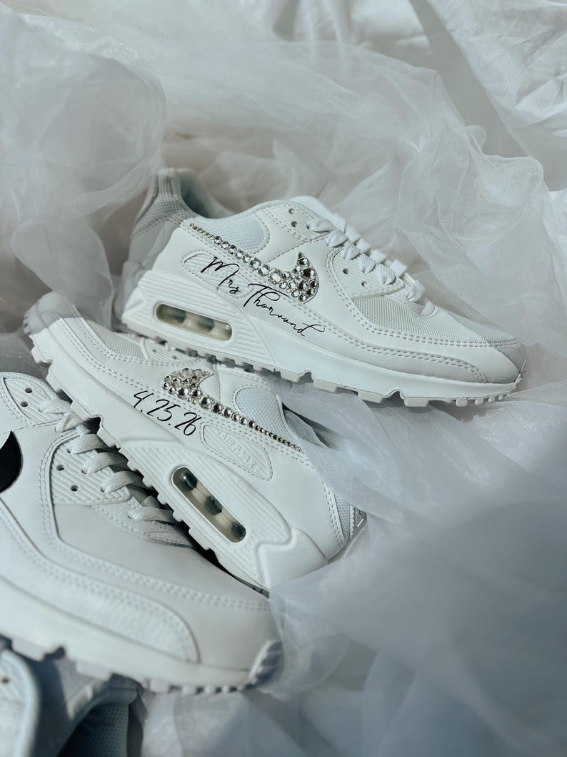 May include: White sneakers with crystal embellishments and the text "Mrs Thorund" and "4.25.26". The sneakers have a visible air pocket in the sole and a black Nike swoosh.